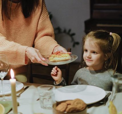 A mother serves dinner to her young daughter at a warmly lit family table, creating a cozy atmosphere.
