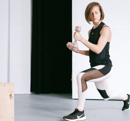 A young woman performing lunges with dumbbells in a modern indoor gym setting.